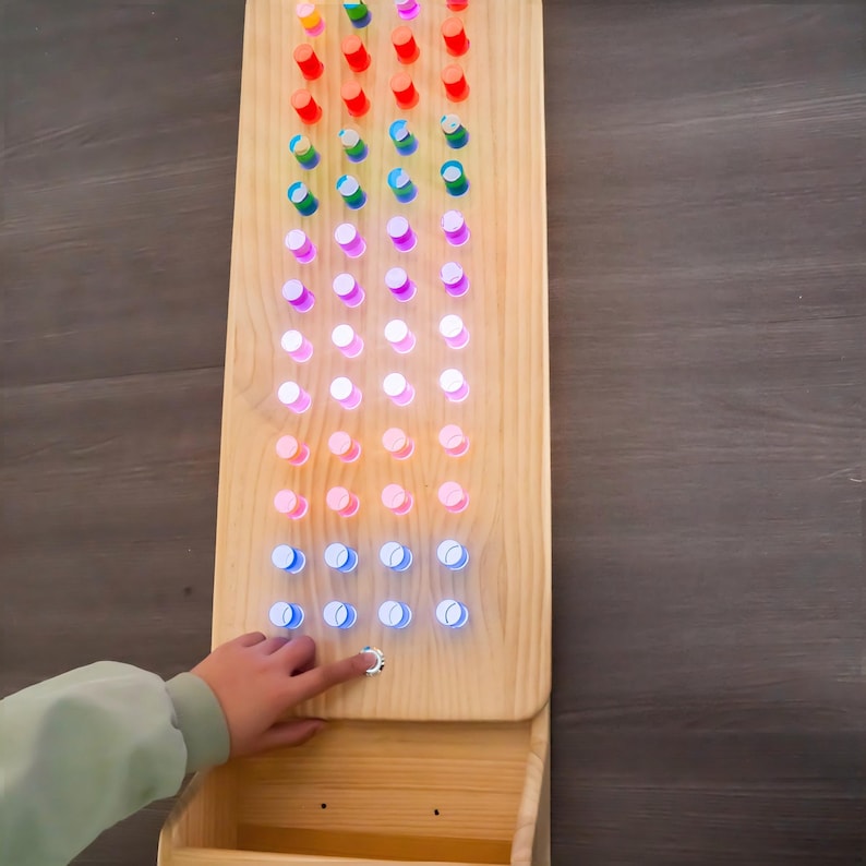 May include: A rectangular light-colored wooden board featuring a grid of circular holes illuminated by internal LED lights in a gradient of colors including red, orange, yellow, green, blue, and purple. A small circular button is located at the bottom.