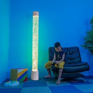 May include: A tall, cylindrical bubble tube lamp with a white base and top. The clear tube is filled with water and small, colorful objects, with bubbles rising. The lamp is lit with a green glow. A child sits nearby.