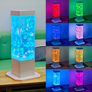 May include: A decorative, rectangular lamp with a white base and top. The lamp's central column is filled with water and colorful, floating fish-shaped ornaments. The lamp is illuminated with various colors, including blue, red, green, and orange.