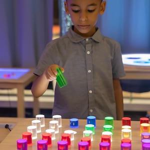 May include: A child holds a green, translucent cylinder above a wooden surface. The surface is covered with an array of illuminated, colorful cylinders in various colors, including white, blue, green, red, and purple. The cylinders appear to be part of an educational toy.