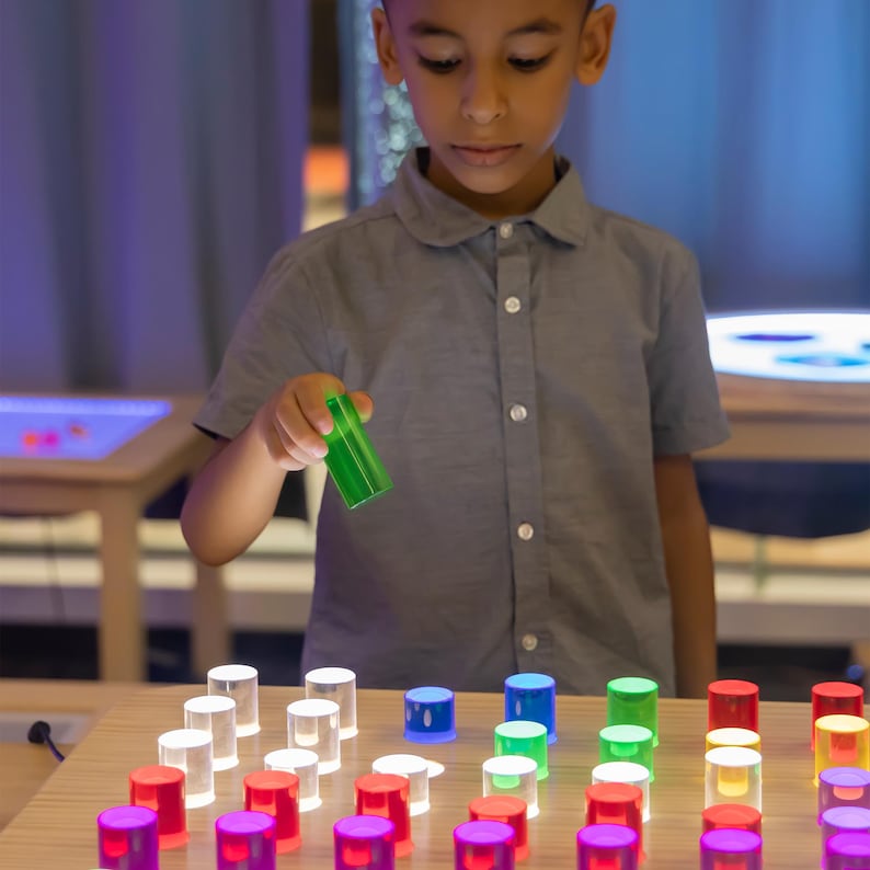 May include: A child holds a green, translucent cylinder above a wooden surface. The surface is covered with an array of illuminated, colorful cylinders in various colors, including white, blue, green, red, and purple. The cylinders appear to be part of an educational toy.