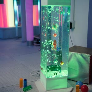May include: A tall, rectangular, illuminated aquarium with colorful fish and bubbles. The aquarium has a white base and top, with a green glow. Several colorful cylinders and spheres are in the foreground.