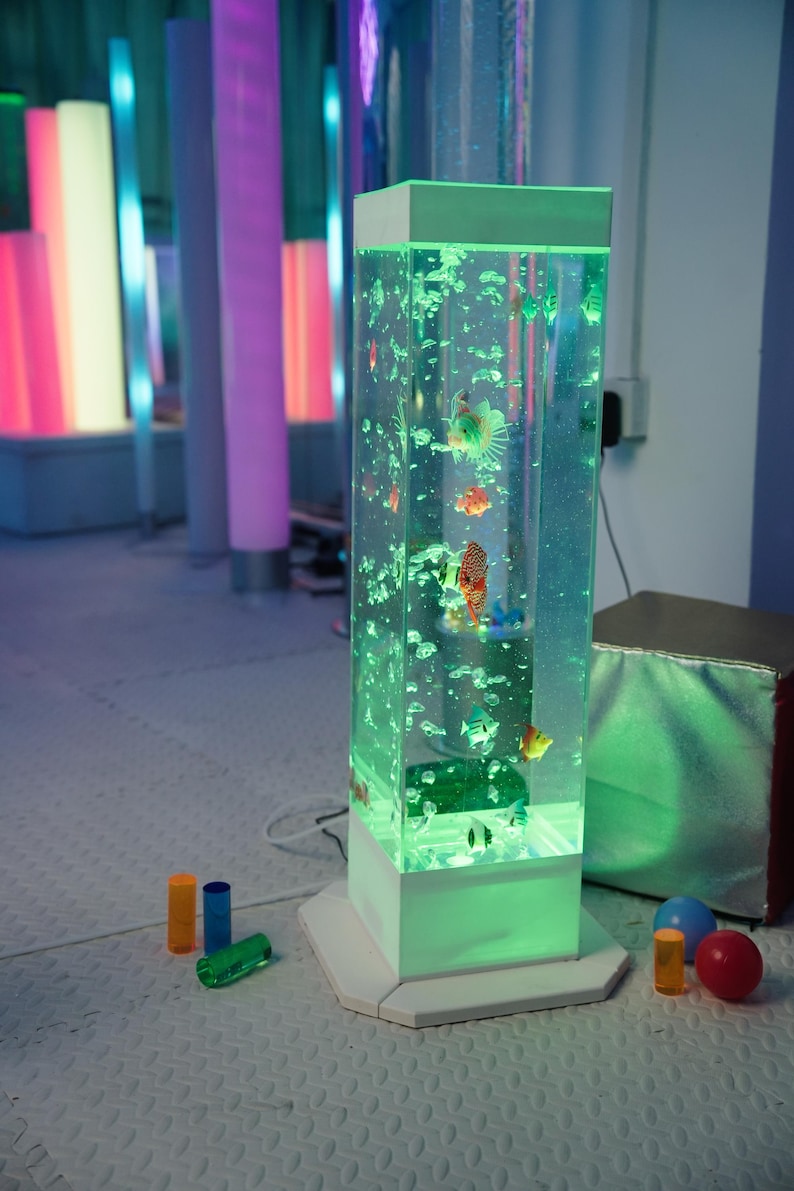 May include: A tall, rectangular, illuminated aquarium with colorful fish and bubbles. The aquarium has a white base and top, with a green glow. Several colorful cylinders and spheres are in the foreground.