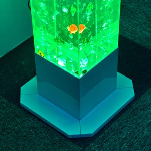 May include: A tall, illuminated aquarium with a green glow, featuring orange and white fish. The aquarium is rectangular with a white base and sits on a matching octagonal platform. The overall design is modern and decorative.