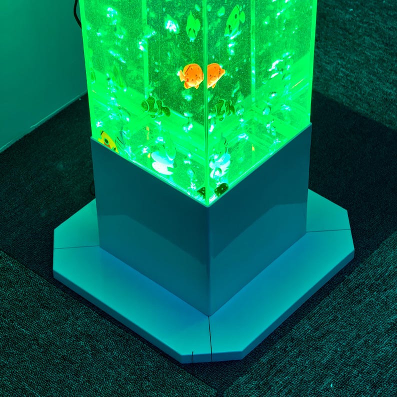 May include: A tall, illuminated aquarium with a green glow, featuring orange and white fish. The aquarium is rectangular with a white base and sits on a matching octagonal platform. The overall design is modern and decorative.