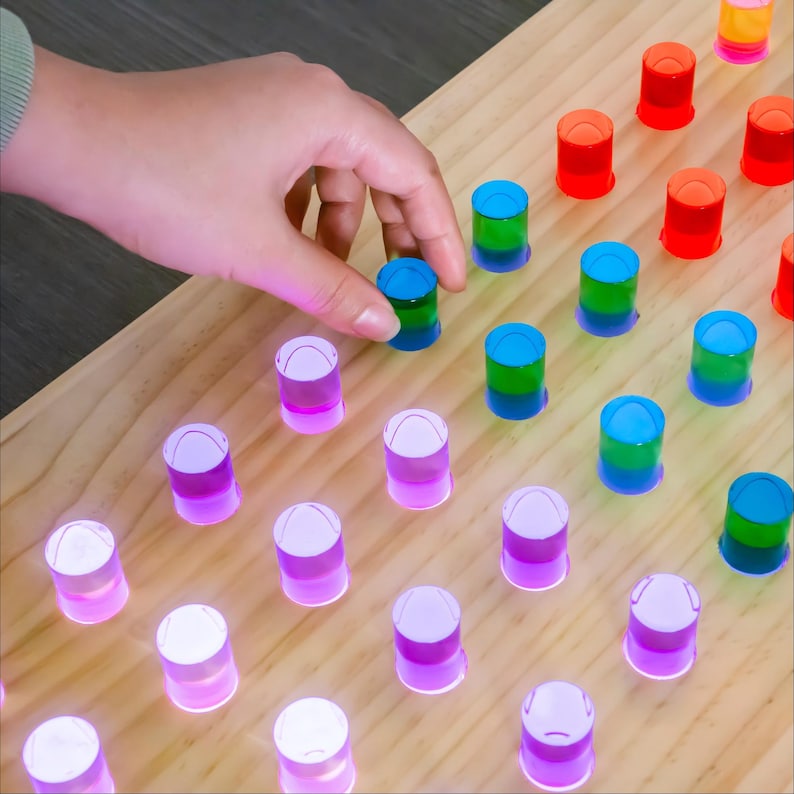 May include: A light-colored wooden board featuring a grid of holes filled with translucent cylindrical pegs in various colors including purple, blue, green, orange, and red. The pegs are arranged in rows and columns, creating a colorful pattern.