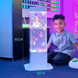 May include: A sensory bubble tube, 80cm tall and 30cm wide, with colorful fish and bubbles. The tube is filled with water and illuminated with lights. The base and top are white. A child is smiling in front of the tube.