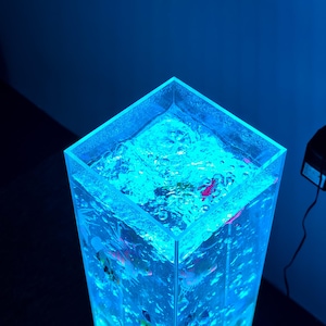 May include: A tall, clear, rectangular aquarium illuminated with blue light. The water inside is bubbling, and small, colorful fish are visible. The aquarium is a decorative piece, likely for home decor or relaxation.