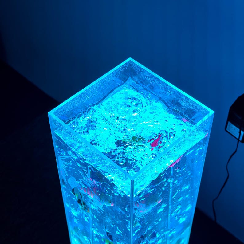 May include: A tall, clear, rectangular aquarium illuminated with blue light. The water inside is bubbling, and small, colorful fish are visible. The aquarium is a decorative piece, likely for home decor or relaxation.