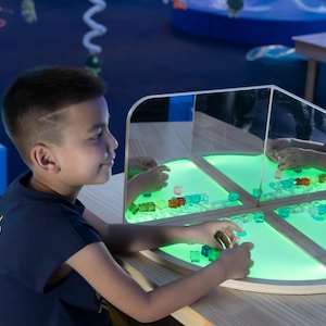 May include: A child interacts with a light-up sensory table. The table has a wooden frame and a mirrored interior, with colorful blocks and a glowing green light. The child is wearing a navy blue shirt.