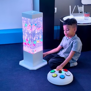 May include: A white, rectangular sensory bubble tube with colorful, floating objects and lights. A child sits on the floor near the tube, interacting with a white, circular control panel with colored buttons. The room has a blue carpet.