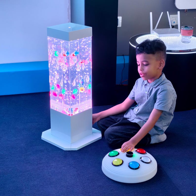 May include: A white, rectangular sensory bubble tube with colorful, floating objects and lights. A child sits on the floor near the tube, interacting with a white, circular control panel with colored buttons. The room has a blue carpet.
