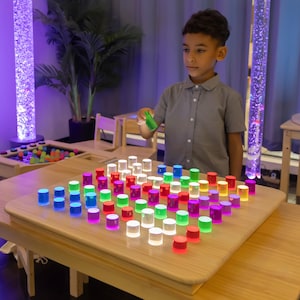 May include: A child interacts with a light-up sensory board featuring a grid of colorful, cylindrical lights. The lights are arranged on a light-colored wooden table. The room has a calming atmosphere with bubble tubes and plants.