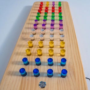 May include: A rectangular light-colored wooden board featuring a grid of forty-eight translucent cylindrical pegs arranged in four columns. The pegs display a gradient of colors including red, yellow, purple, green, silver, gold, and blue. A single circular metal button is centered at the bottom.