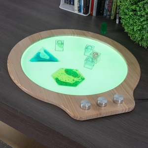 May include: A wooden-framed light table with a glowing green surface. The table features a variety of colorful, geometric shapes, including a triangle and a hexagon. Three silver knobs are located on the lower right side of the table.