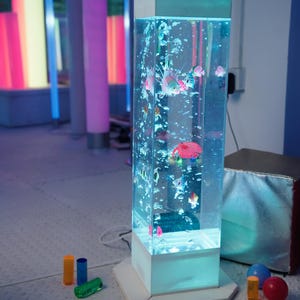 May include: A rectangular, illuminated aquarium with colorful, artificial fish and bubbles. The tank is white-based and sits on a white surface. Colorful, cylindrical lights are in the background, along with small, colorful toys.