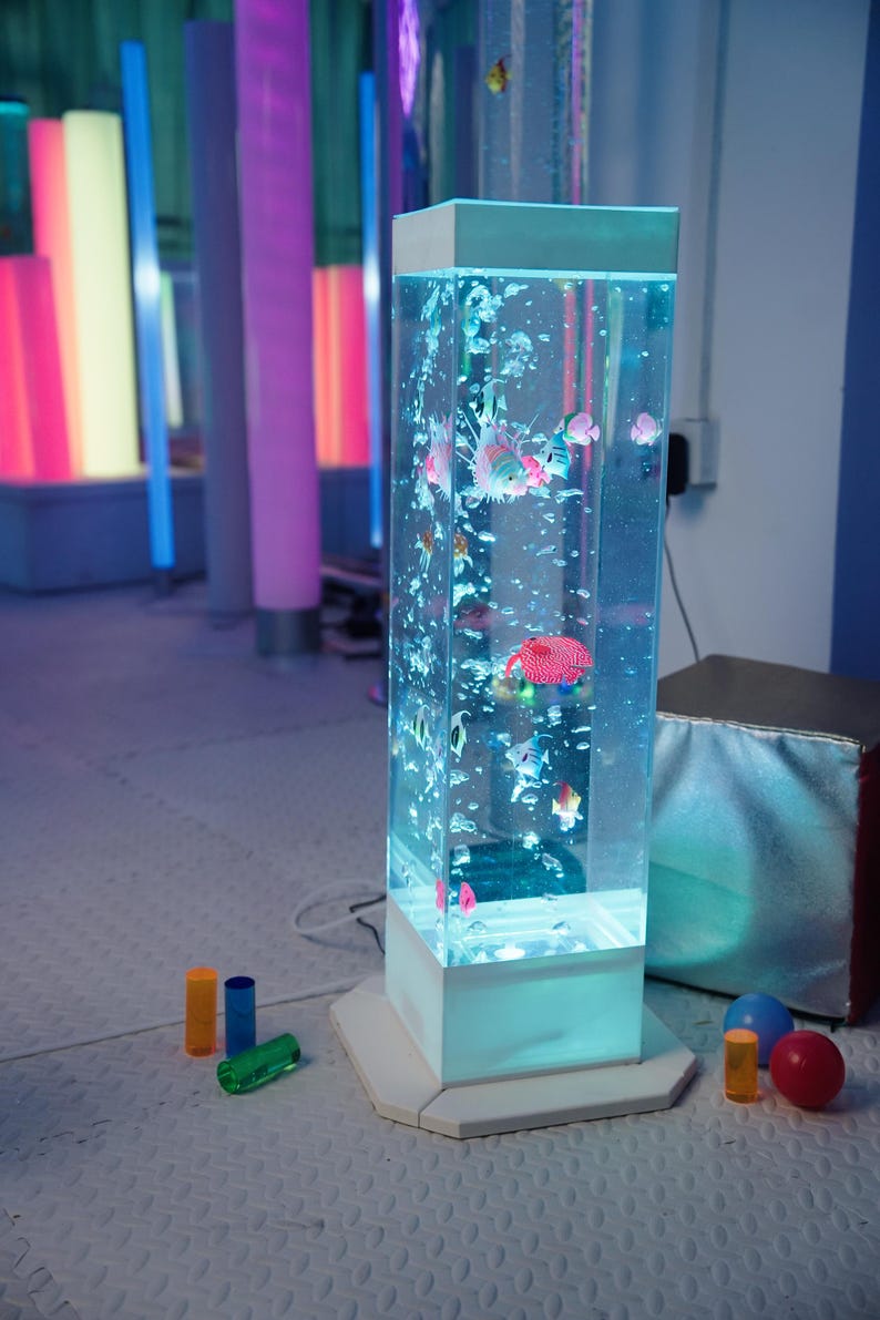 May include: A rectangular, illuminated aquarium with colorful, artificial fish and bubbles. The tank is white-based and sits on a white surface. Colorful, cylindrical lights are in the background, along with small, colorful toys.