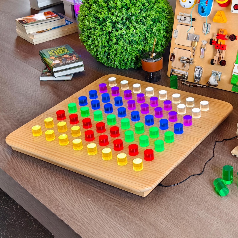 May include: A wooden pegboard game with rows of colorful pegs in yellow, red, green, blue, purple, and white. The board is on a wooden table, with other toys and books in the background.