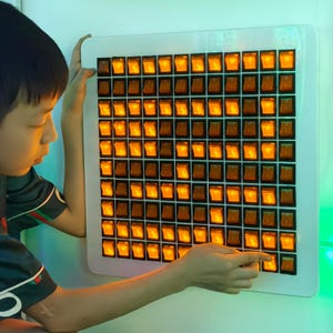 May include: A white square panel with a grid of orange illuminated toggle switches. The switches are arranged in rows and columns. A person is interacting with the switches, pointing at one with their finger. The switches are lit up, creating a warm glow.