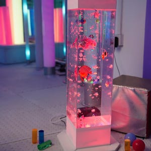 May include: A tall, rectangular, clear aquarium with a white base and top, illuminated with pink light. The aquarium contains colorful, artificial fish and bubbles. Several colorful cylindrical and spherical objects are in the foreground.