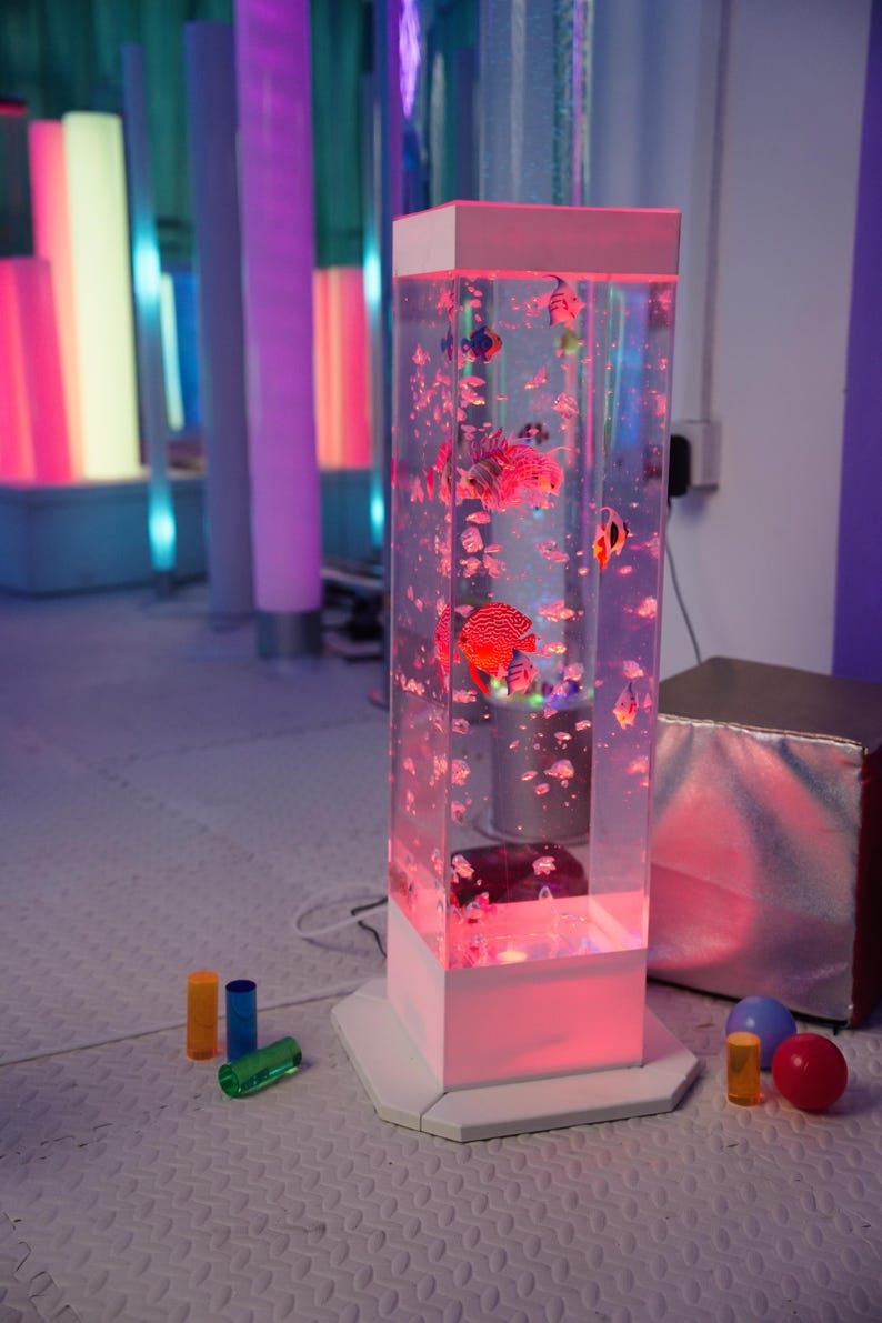 May include: A tall, rectangular, clear aquarium with a white base and top, illuminated with pink light. The aquarium contains colorful, artificial fish and bubbles. Several colorful cylindrical and spherical objects are in the foreground.