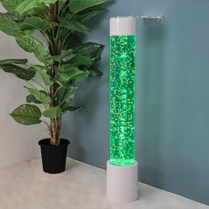 May include: A tall, cylindrical sensory bubble tube lamp with a white base and top. The tube is filled with green-tinted water and floating yellow and orange bubbles. The lamp is next to a large potted plant with green leaves.
