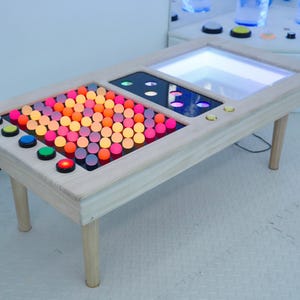 May include: A wooden table with a light-colored frame and four legs. The table's surface features illuminated sections with colorful, round lights and buttons. The table has a modern, interactive design.
