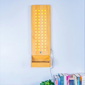 May include: A vertical rectangular light fixture made of light-toned wood featuring a grid of circular cutouts that emit a warm yellow glow. The fixture includes a small integrated shelf at the bottom and a black power cord extending to the side.