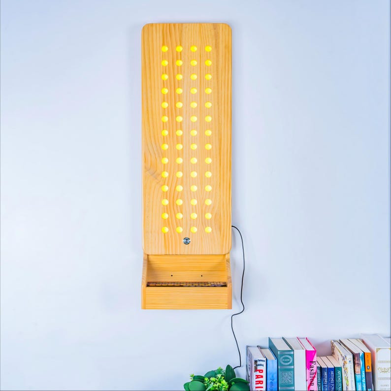 May include: A vertical rectangular light fixture made of light-toned wood featuring a grid of circular cutouts that emit a warm yellow glow. The fixture includes a small integrated shelf at the bottom and a black power cord extending to the side.