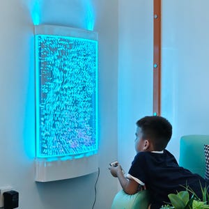 May include: A wall-mounted bubble panel with blue LED lighting. The panel is rectangular with a curved top and bottom, and the bubbles are illuminated. A child is holding a remote control and looking at the panel.