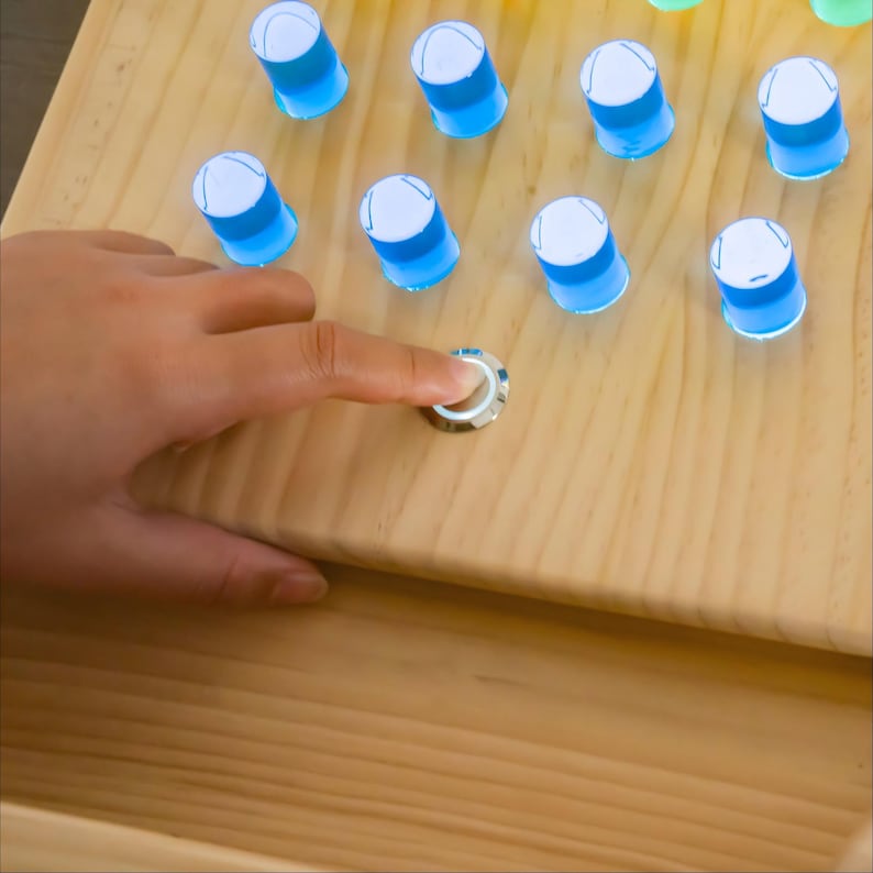 May include: A light-colored wooden board featuring multiple translucent blue cylindrical pegs arranged in a grid. A circular silver metal button is embedded in the wood surface, which is being pressed to activate the light-up function of the pegs.