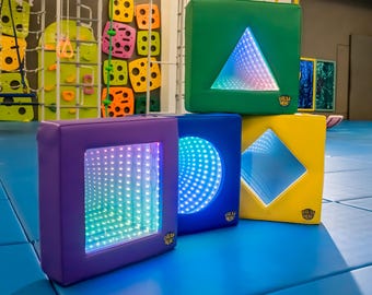 Puede incluir: Cuatro bloques sensoriales de colores con formas geométricas. Los bloques son morado, azul, verde y amarillo. Cada bloque tiene una forma diferente recortada, con un interior espejado y luces LED. El bloque verde tiene un triángulo, el azul un círculo y el amarillo un rombo.