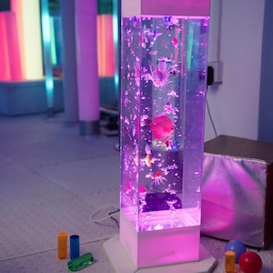 May include: A tall, rectangular bubble tube with a white base and top, illuminated with pink and purple lights. The tube contains colorful, plastic fish and bubbles. Small, colorful cylinders and spheres are scattered on the floor.