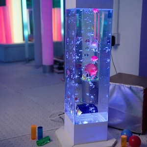 May include: A tall, rectangular bubble tube with a white base and top. The tube is filled with water, colorful fish, and bubbles. The tube is illuminated with blue light. Colorful cylinders and spheres are in the foreground.