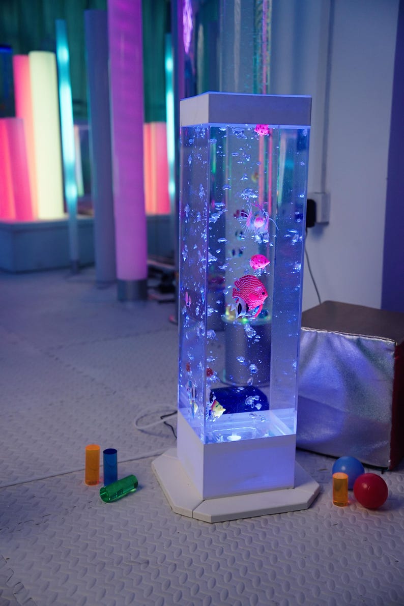 May include: A tall, rectangular bubble tube with a white base and top. The tube is filled with water, colorful fish, and bubbles. The tube is illuminated with blue light. Colorful cylinders and spheres are in the foreground.