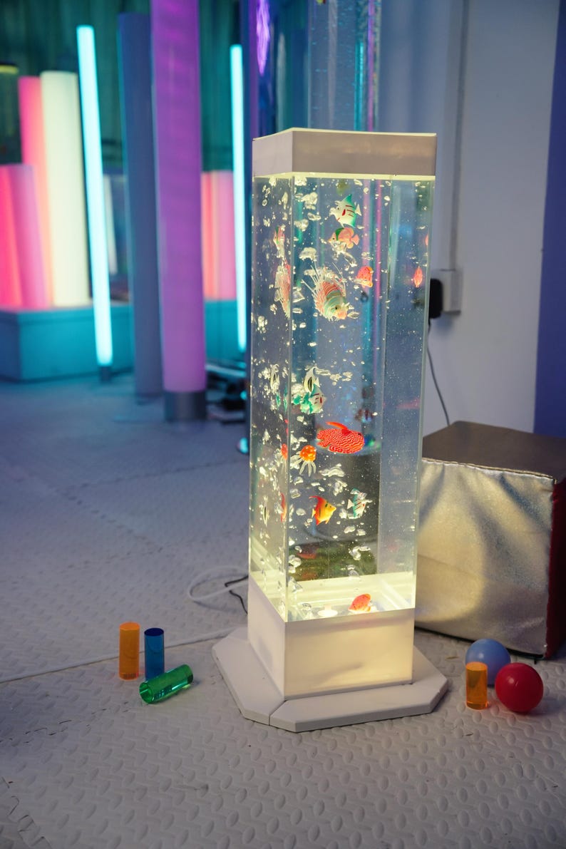 May include: A tall, rectangular aquarium lamp with colorful, animated fish. The lamp has a white base and top, with a clear, water-filled column. Bubbles and various toy fish float inside. Additional colorful cylinders and spheres are in the foreground.