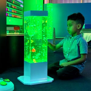 May include: A tall, rectangular sensory bubble tube lamp with a white base and top. The lamp is filled with green liquid and floating colorful shapes. A young person is interacting with the lamp in a brightly lit room.