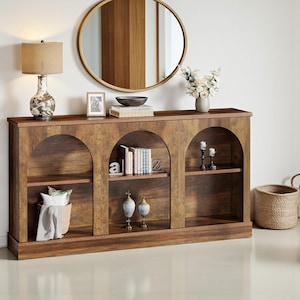 May include: A wooden bookshelf with three arched openings. The bookshelf is a warm brown colour and has shelves for storage. A lamp, mirror, and decorative items are on top of the bookshelf. The room is decorated in a neutral colour palette.