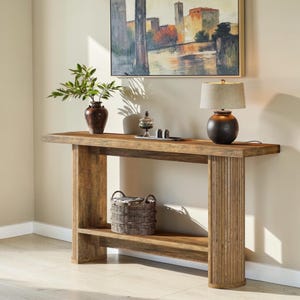 May include: A wooden console table with a lower shelf and ribbed supports. A woven basket sits on the shelf. The table is topped with a lamp, a vase with greenery, and decorative items. An abstract painting hangs above.