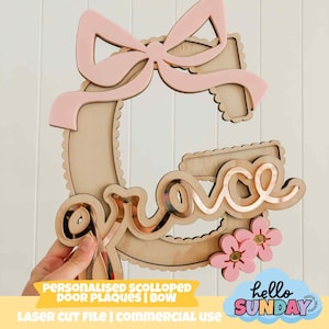 以下が含まれることがあります： 「G」の形をした木製のドアプレート。名前「Grace」はミラー仕上げ。ピンクのリボンと2つのピンクの花が特徴です。「PERSONALIZED SCOLLOPED DOOR PLAQUES | BOW」の文字が見えます。