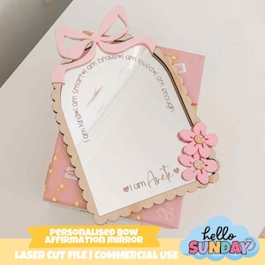 May include: A personalized affirmation mirror with a bow and flower design. The mirror has a scalloped edge and features the phrases "I am kind, smart, brave, loved, enough." The text "I am Arete" is also present. The mirror is pink and light brown.