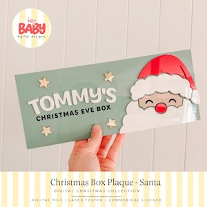 May include: A rectangular sage green plaque featuring a layered Santa Claus face on the right side with a red hat and white beard. The text reads TOMMY'S CHRISTMAS EVE BOX in white and black lettering, accented by four small gold stars.