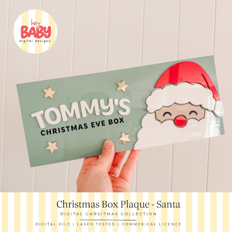 May include: A rectangular sage green plaque featuring a layered Santa Claus face on the right side with a red hat and white beard. The text reads TOMMY'S CHRISTMAS EVE BOX in white and black lettering, accented by four small gold stars.