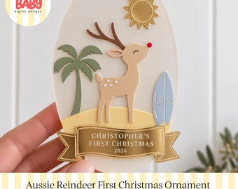 Könnte beinhalten: Ein ovaler Anhänger mit einem beigen Rentier mit roter Nase, einer grünen Palme, einem hellblauen Surfbrett und einer goldenen Sonne. Ein goldenes Banner unten zeigt den Text: CHRISTOPHER'S FIRST CHRISTMAS 2026.