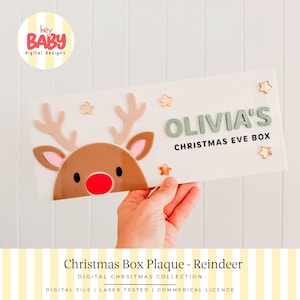 May include: A rectangular white plaque featuring a brown reindeer face with pink inner ears and a bright red nose. The text reads OLIVIA'S CHRISTMAS EVE BOX in green and black lettering, accented by small gold stars scattered across the surface.