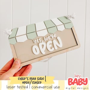 May include: Wooden sign with the text "YES, WE'RE OPEN" in white, with a light green and white awning design at the top. The sign is rectangular with a light brown frame and is designed for a child's play area.