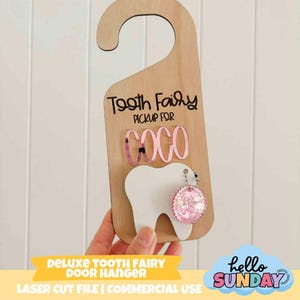 以下が含まれることがあります： 上部が湾曲した木製のドアハンガーで、「Tooth Fairy PICKUP FOR COCO」の文字が入っています。白い歯の形とピンクのチャームが付いています。下のバナーには「DELUXE TOOTH FAIRY DOOR HANGER」と書かれています。