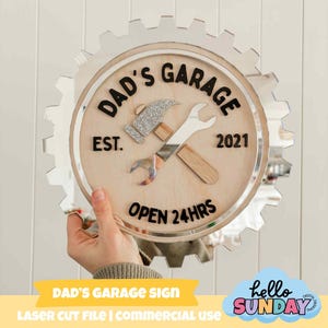 May include: A round wooden sign with the text "DAD'S GARAGE" and "EST. 2021" in black. The sign features a silver hammer and wrench design. The sign is surrounded by a gear-shaped border and includes the text "OPEN 24HRS".