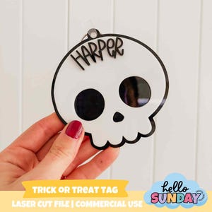 May include: A white skull-shaped tag with black outlines and eye sockets, the name "HARPER" is written across the top. The tag is held by a person. The words "TRICK OR TREAT TAG" are printed on a yellow banner.