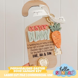 May include: Wooden Easter door hanger key with the words "Easter Bunny" in pink and green. It features a carrot and a ribbon. The text reads "Personalised Easter Door Hanger Key".